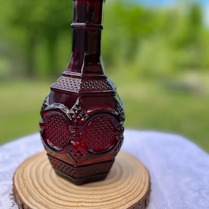 Peut inclure: Une carafe en verre rouge profond présentant un motif géométrique texturé avec des formes en diamant et des motifs circulaires. La bouteille possède un col haut et fin ainsi qu'un corps large et arrondi, fabriquée en verre pressé coloré.