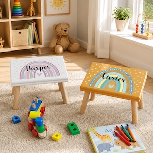 May include: Two wooden step stools with four legs each. One stool is white with a pastel rainbow and the name Harper. The other stool is mustard yellow with a blue and yellow rainbow and the name Carter. Both feature small heart patterns.
