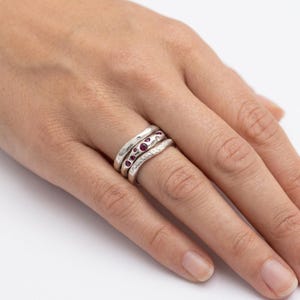 Op de afbeelding: Een stapel van drie zilveren ringen gedragen aan een vinger. De bovenste ring is glad en effen. De middelste ring bevat een reeks kleine, ingebedde ronde roze edelstenen. De onderste ring heeft een getextureerd, korrelig oppervlak aan de rand.