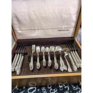 Può includere: Una cassetta per posate in legno con rivestimento interno color crema recante la scritta DEEPSILVER. Il set comprende coltelli, forchette, cucchiai e un mestolo color argento disposti in un vassoio foderato in velluto marrone.