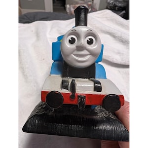 May include: A blue and black toy train engine featuring a grey face with large eyes and a smiling mouth. The train has a black chimney, a red front bumper with two black buffers, and is mounted on a black textured base representing train tracks.