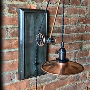 K&ouml;nnte beinhalten: Eine Wandleuchte im Steampunk-Stil aus Kupfer und Schwarz mit einem Flaschenzugsystem und einem Kupferschirm. Die Leuchte ist an einer dunkelgr&uuml;n lackierten Holzr&uuml;ckwand montiert.