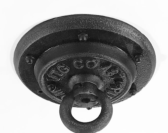 May include: A black, industrial-style ceiling canopy with a circular design. It features a central ring for hanging and is secured with multiple bolts. The surface has a textured, cast-iron appearance, with the text "TG CO" visible.