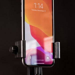 Pode incluir: Um smartphone preto com uma tela branca que mostra a hora 1:14 e a data ter&ccedil;a-feira, 17 de novembro. O telefone est&aacute; montado em um suporte de telefone preto com uma manivela no lado. O telefone est&aacute; conectado a um cabo preto.