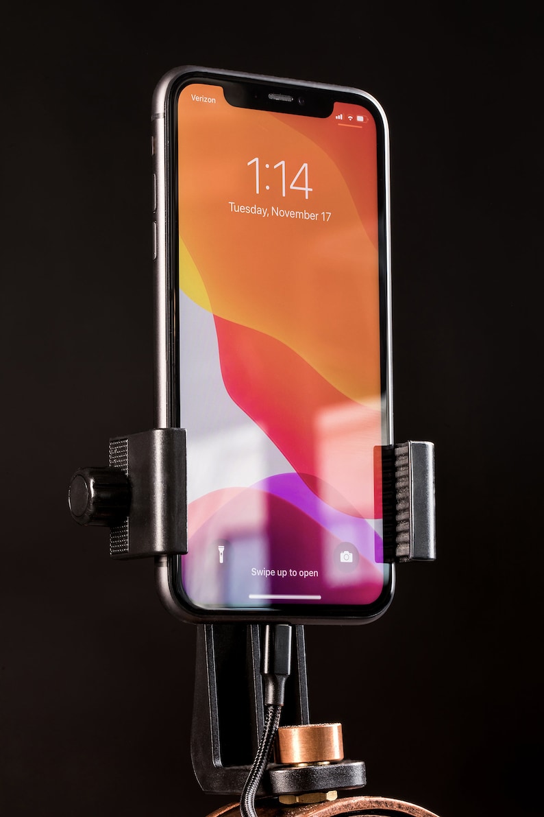 Pode incluir: Um smartphone preto com uma tela branca que mostra a hora 1:14 e a data ter&ccedil;a-feira, 17 de novembro. O telefone est&aacute; montado em um suporte de telefone preto com uma manivela no lado. O telefone est&aacute; conectado a um cabo preto.