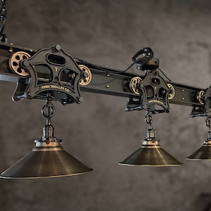 May include: A black industrial-style ceiling light fixture with three hanging shades. The fixture is made of metal and has a vintage look. The shades are made of metal and have a brass finish. The fixture is labeled "Ohio Trolley Co."