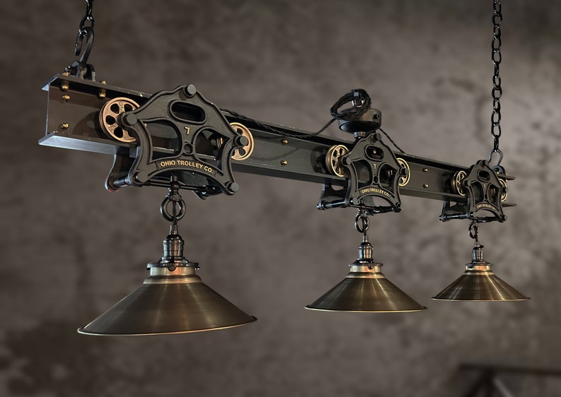 May include: A black industrial-style ceiling light fixture with three hanging shades. The fixture is made of metal and has a vintage look. The shades are made of metal and have a brass finish. The fixture is labeled "Ohio Trolley Co."