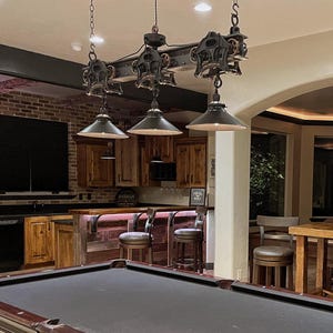 May include: A black industrial-style light fixture with three pendant lights hangs over a pool table in a game room. The light fixture is made of metal and has a rustic look. The lights are hanging from a track system with pulleys and gears. The pool table is green felt and has a wooden frame. The room has a bar area with wooden stools and a brick wall.