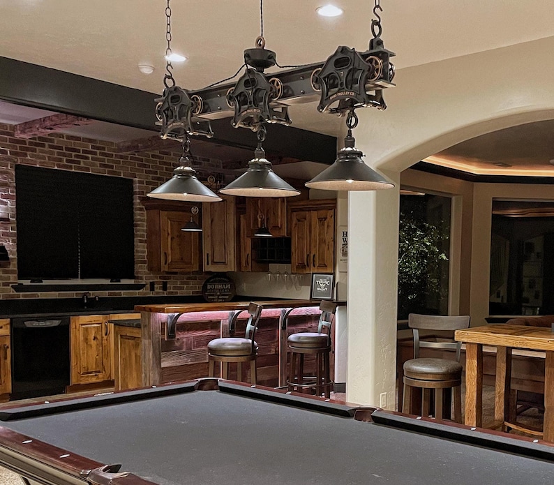 May include: A black industrial-style light fixture with three pendant lights hangs over a pool table in a game room. The light fixture is made of metal and has a rustic look. The lights are hanging from a track system with pulleys and gears. The pool table is green felt and has a wooden frame. The room has a bar area with wooden stools and a brick wall.