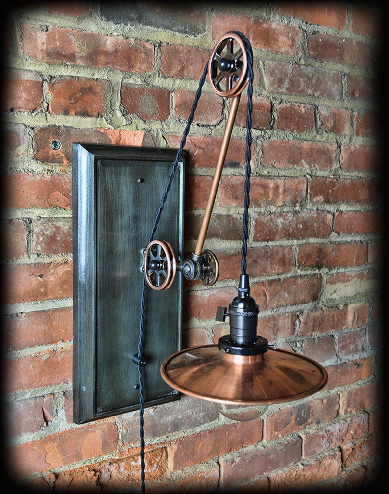 K&ouml;nnte beinhalten: Eine Wandleuchte im Steampunk-Stil aus Kupfer und Schwarz mit einem Flaschenzugsystem und einem Kupferschirm. Die Leuchte ist an einer dunkelgr&uuml;n lackierten Holzr&uuml;ckwand montiert.