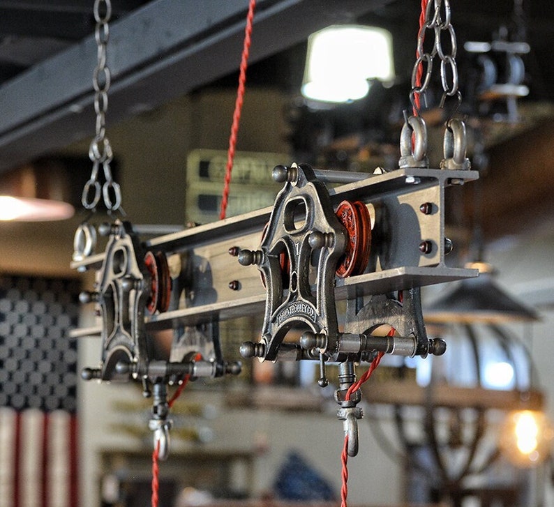 May include: A vintage industrial style light fixture with a metal frame and red rope cords. The fixture has two sets of pulleys and is suspended from the ceiling by chains.