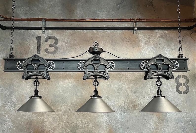 May include: Three industrial-style pendant lights with black metal shades hanging from a black metal beam with three black metal pulleys. The beam is attached to the ceiling with chains. The pulleys have the text "Ohio Trolley Co." on them. The numbers "13" and "8" are painted on the wall behind the lights.