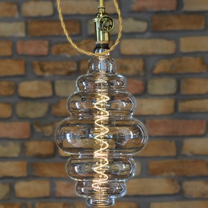 May include: A clear glass pendant light bulb with a coiled filament and a brass hanging fixture. The bulb has a unique, layered design with multiple rounded sections.
