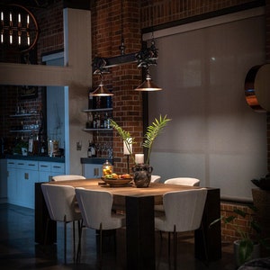 May include: A wooden dining table with six white chairs, set in a loft-style dining room with exposed brick walls and a large window with a white blind. The table is decorated with a vase of green plants, a bowl of fruit, and a candle.