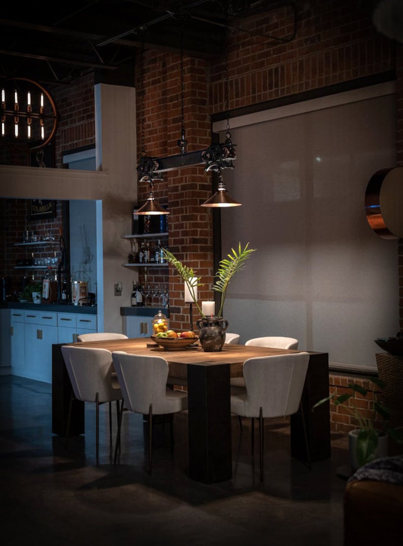 May include: A wooden dining table with six white chairs, set in a loft-style dining room with exposed brick walls and a large window with a white blind. The table is decorated with a vase of green plants, a bowl of fruit, and a candle.