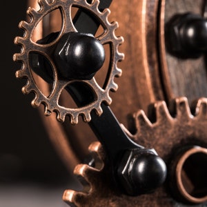 Pode incluir: Close-up de um conjunto de engrenagens de bronze interligadas com detalhes pretos. As engrenagens s&atilde;o conectadas por uma haste de metal preta.