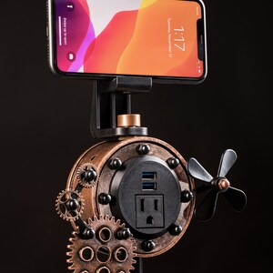 Pode incluir: Um suporte para telefone de estilo steampunk com acabamento em cobre e preto. O suporte possui uma tomada de energia e uma porta USB integradas, bem como um ventilador rotativo. Um smartphone est&aacute; montado no suporte, mostrando a hora e a data.