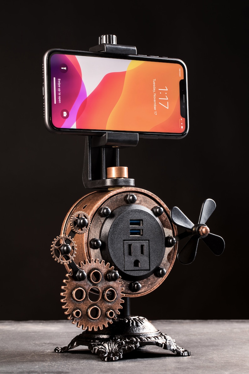 Pode incluir: Um suporte para telefone de estilo steampunk com acabamento em cobre e preto. O suporte possui uma tomada de energia e uma porta USB integradas, bem como um ventilador rotativo. Um smartphone est&aacute; montado no suporte, mostrando a hora e a data.
