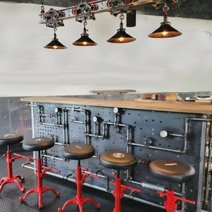 May include: Four black industrial-style pendant lights with metal shades hang from a track system over a bar counter with red metal stools. The counter has a black metal backsplash with a grid pattern and pipes.