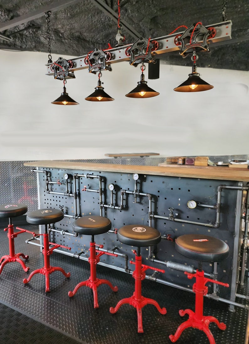 May include: Four black industrial-style pendant lights with metal shades hang from a track system over a bar counter with red metal stools. The counter has a black metal backsplash with a grid pattern and pipes.