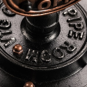 May include: Close-up of a black metal pipe fitting with the words "ROOM PIPE" embossed on the side. The fitting has a copper-colored cap on top and is decorated with copper-colored rivets.