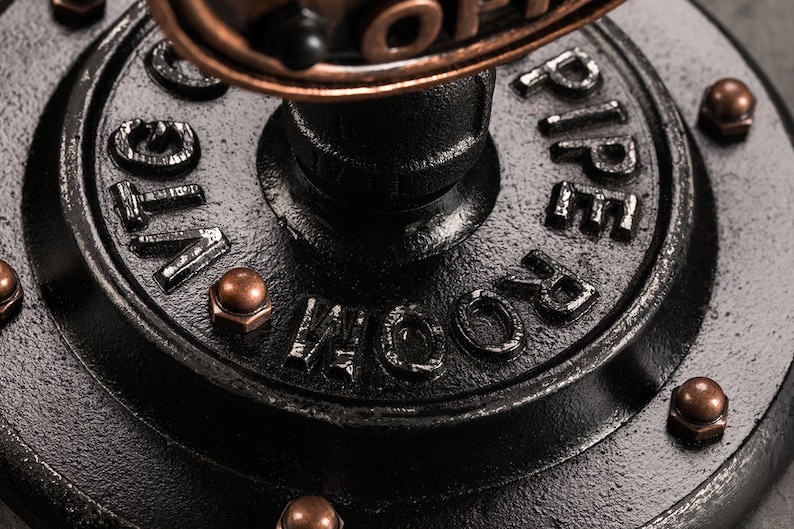 May include: Close-up of a black metal pipe fitting with the words "ROOM PIPE" embossed on the side. The fitting has a copper-colored cap on top and is decorated with copper-colored rivets.