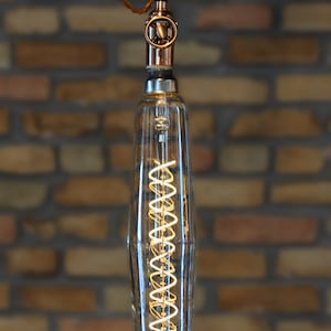May include: A clear glass light bulb with a copper colored metal top and a coiled filament inside. The bulb is hanging from a brown cord against a brick wall background.