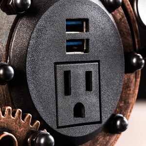 Pode incluir: Close-up de uma tomada el&eacute;trica preta com duas portas USB. A tomada &eacute; cercada por uma moldura de metal de cor cobre com engrenagens e rebites.