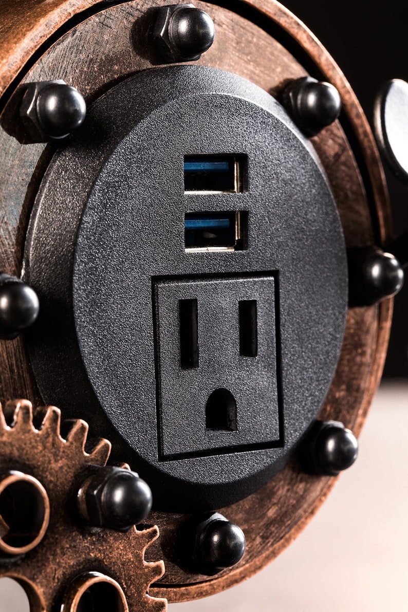 Pode incluir: Close-up de uma tomada el&eacute;trica preta com duas portas USB. A tomada &eacute; cercada por uma moldura de metal de cor cobre com engrenagens e rebites.
