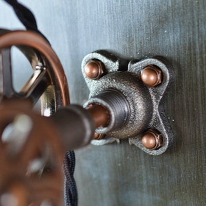 K&ouml;nnte beinhalten: Nahaufnahme einer Metallrohrverbindung mit vier kupferfarbenen Muttern. Die Verbindung ist an einer Wand befestigt und hat ein Gewindeende.