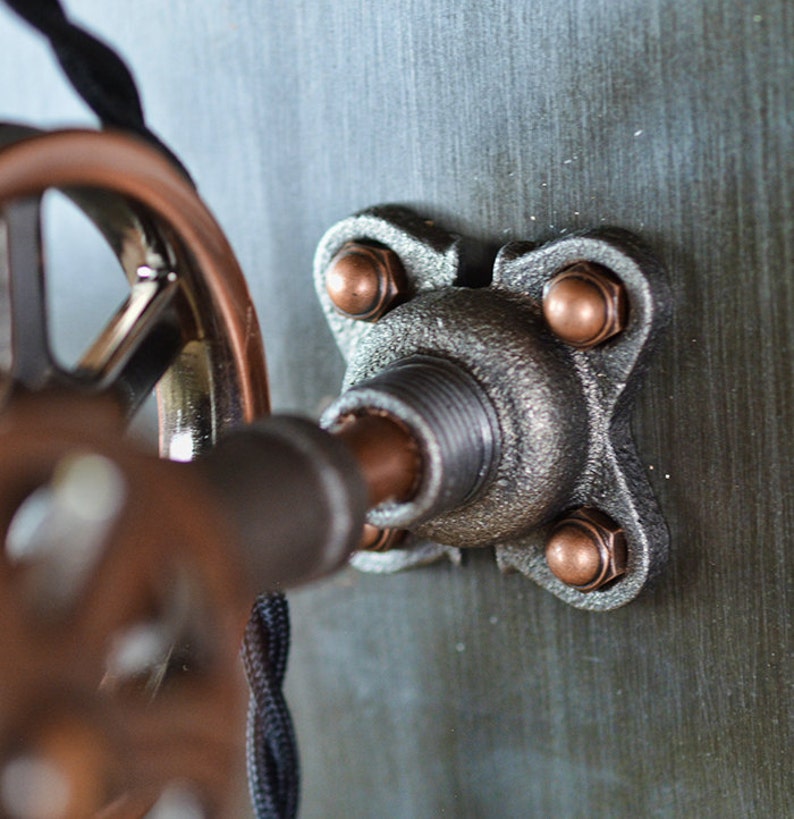 K&ouml;nnte beinhalten: Nahaufnahme einer Metallrohrverbindung mit vier kupferfarbenen Muttern. Die Verbindung ist an einer Wand befestigt und hat ein Gewindeende.