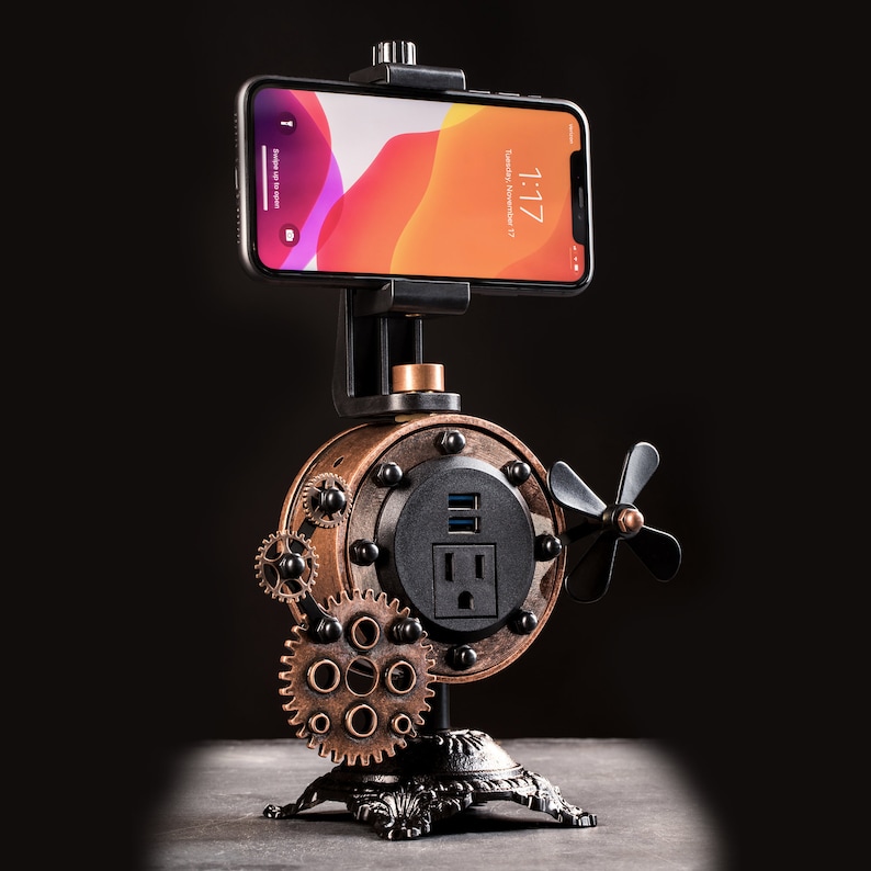 Pode incluir: Um suporte para telefone estilo steampunk com acabamento preto e cobre. Possui um suporte para telefone, uma tomada de energia e portas USB. O suporte est&aacute; sobre uma superf&iacute;cie preta.