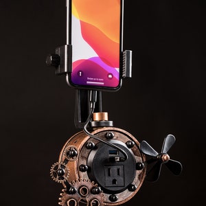 Pode incluir: Uma esta&ccedil;&atilde;o de carregamento de telefone estilo steampunk com acabamento preto e cobre. Possui uma tomada de energia integrada, uma porta USB e um suporte para telefone. A esta&ccedil;&atilde;o &eacute; montada em uma base de metal preto com engrenagens decorativas.