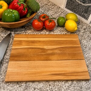 Puede incluir: Una tabla de cortar rectangular de madera con veta natural se encuentra junto a un cuchillo de chef de acero inoxidable con mango negro. Cerca hay un cuenco de madera con pimientos y brócoli, dos tomates rojos y dos limones amarillos.