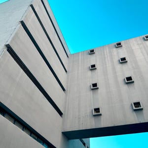 Può includere: Una vista dal basso di un edificio in cemento grigio con una struttura a ponte che collega due sezioni. La facciata è caratterizzata da linee pulite, fasce orizzontali rientrate e una griglia di piccole finestre quadrate profondamente incassate su un cielo azzurro brillante.