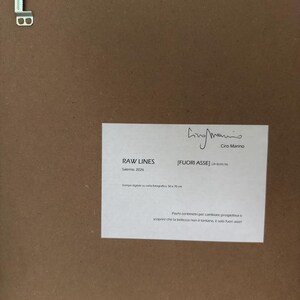 Op de afbeelding: De achterkant van een ingelijst kunstwerk met een bruine achterplaat en een metalen ophangbeugel. Een wit label toont de tekst RAW LINES, FUORI ASSE, Ciro Marino, Salerno 2026, Stampa digitale su carta fotografica, 50x70 cm, en tekst.
