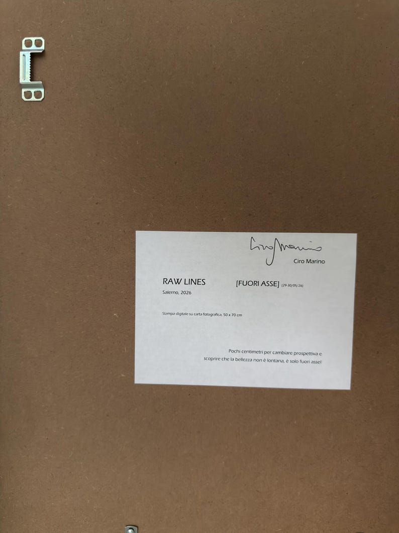 Op de afbeelding: De achterkant van een ingelijst kunstwerk met een bruine achterplaat en een metalen ophangbeugel. Een wit label toont de tekst RAW LINES, FUORI ASSE, Ciro Marino, Salerno 2026, Stampa digitale su carta fotografica, 50x70 cm, en tekst.