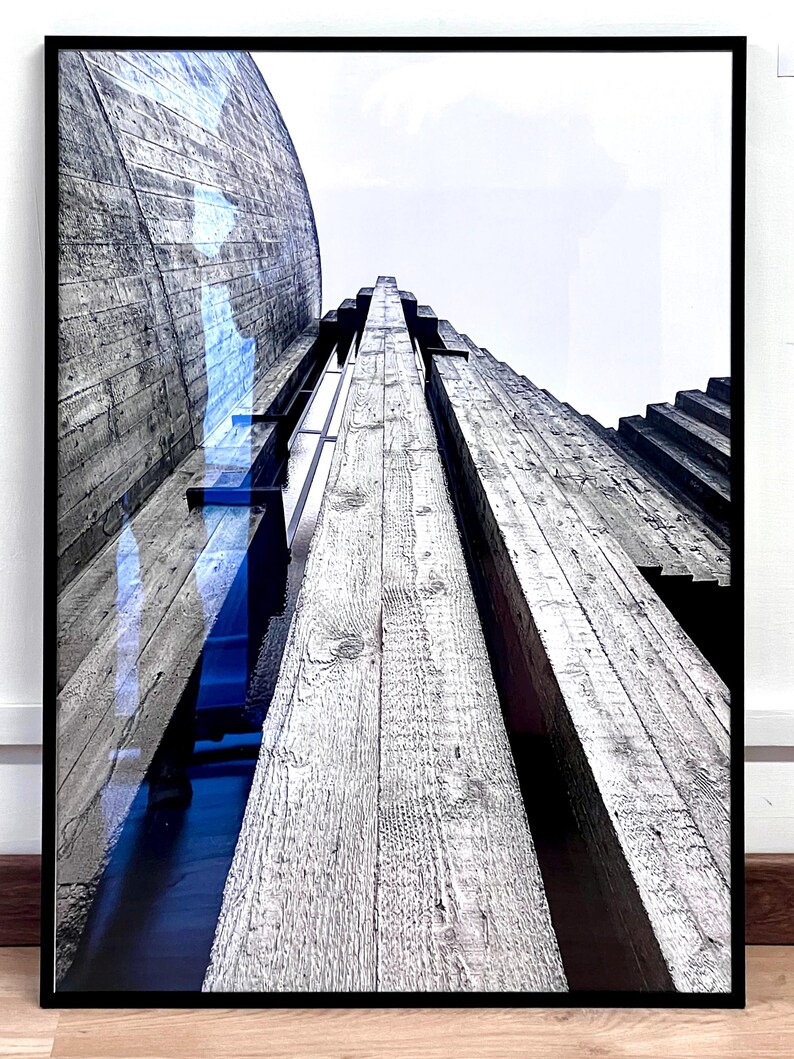 Op de afbeelding: Een ingelijste wandkunstprint met een kikvorsperspectief van een betonstructuur met opvallende houtnerftexturen. De compositie bevat verticale lijnen, een gebogen wandgedeelte en een diepblauwe reflectie op het glazen oppervlak.