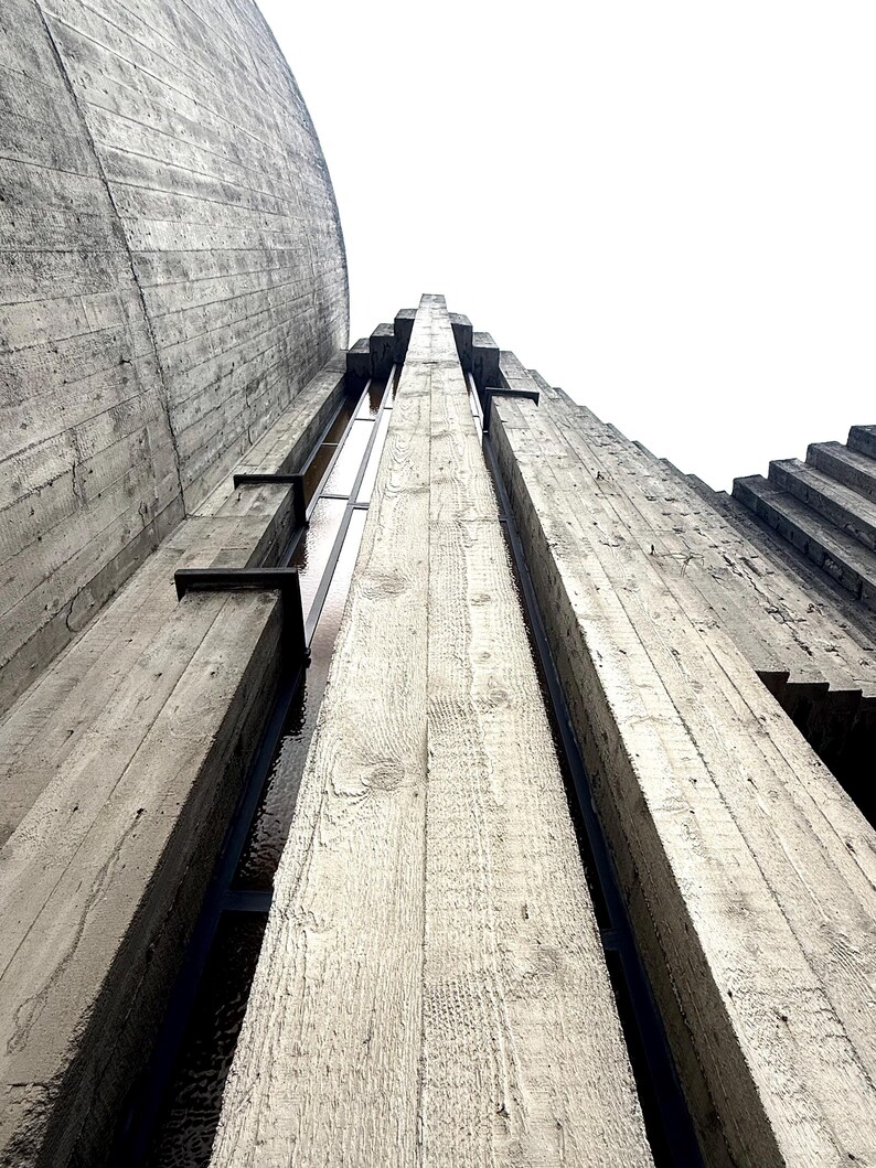Op de afbeelding: Een kikvorsperspectief van een hoge grijze betonstructuur met verticale, getextureerde zuilen met zichtbare houtnerfafdrukken. Het ontwerp bevat smalle, verzonken verticale ramen tussen de betonnen kolommen die omhoog reiken naar een heldere lucht.