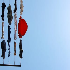 Può includere: Un'installazione mobile verticale caratterizzata da molteplici fili metallici sottili sospesi tra due barre orizzontali. Vari pezzi di tessuto in nero, blu, bianco e un prominente tessuto circolare rosso sono fissati ai fili, creando una disposizione a strati.