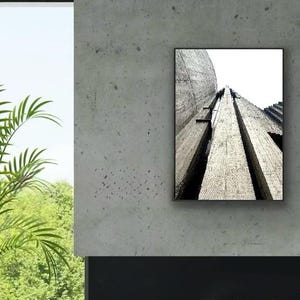 Op de afbeelding: Een rechthoekige wandkunstprint met een zwart-wit architectuurfoto van een gestructureerde betonconstructie. De afbeelding toont een kikkerperspectief van verticale betonzuilen en een gebogen muur, wat diepe schaduwen en lijnen cre&euml;ert.