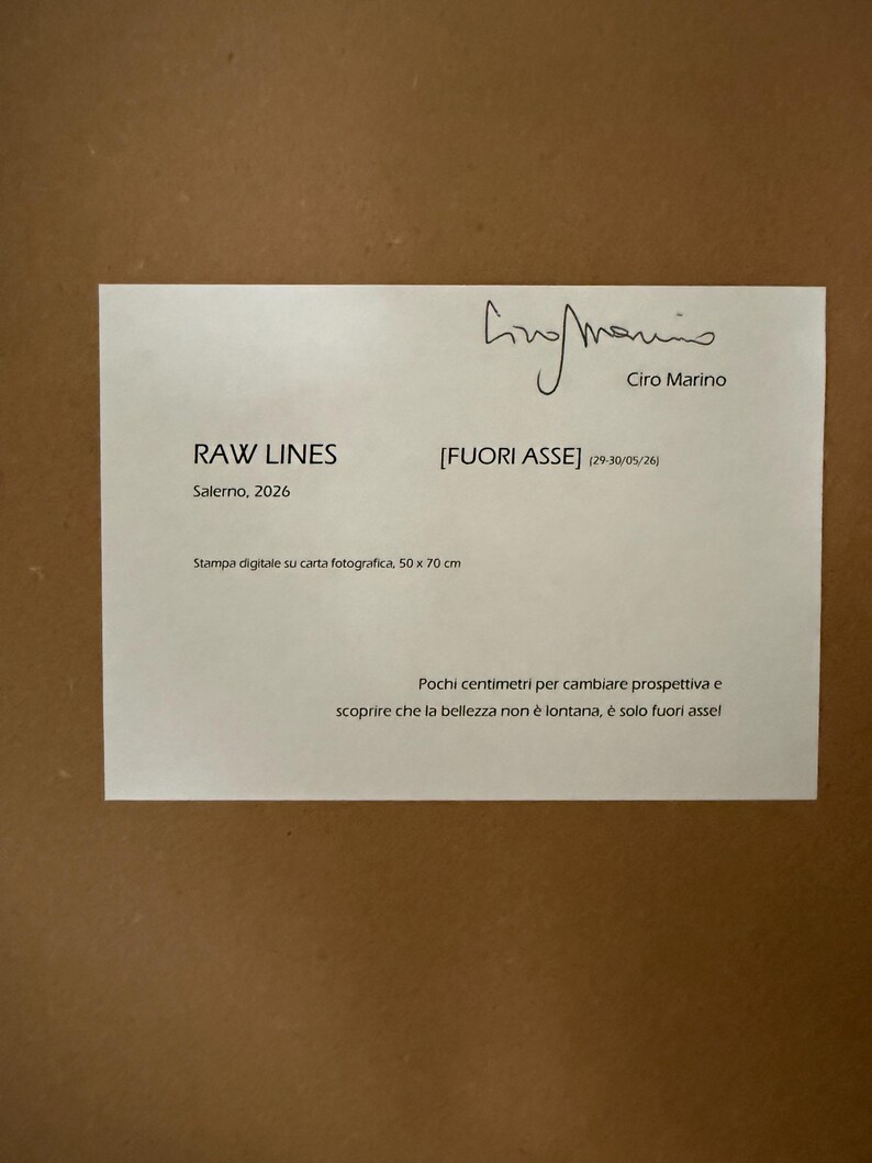 Op de afbeelding: Een witte rechthoekige informatiekaart met zwarte tekst, voorzien van de handtekening van Ciro Marino, de titel RAW LINES [FUORI ASSE], de datum 2026, de tekst Stampa digitale su carta fotografica, 50 x 70 cm en een korte Italiaanse zin.
