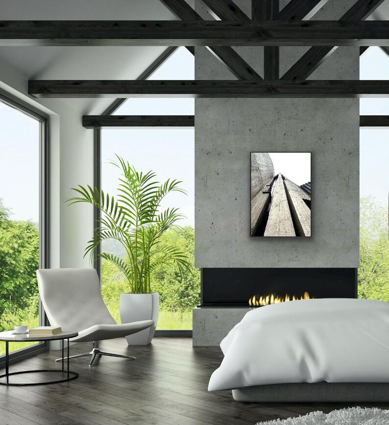Op de afbeelding: Een rechthoekige wandkunstprint met een zwart-wit architectuurfoto vanuit een laag standpunt van gestructureerde betonconstructies met diepe verticale groeven, ingelijst in een dunne zwarte lijst aan een grijze betonnen muur.