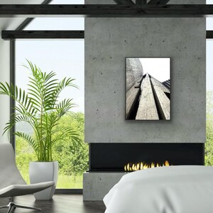 Op de afbeelding: Een rechthoekige wandkunstprint met een zwart-wit architectuurfoto van hoge, getextureerde betonstructuren die omhoog reiken naar een heldere lucht, weergegeven in een dunne zwarte lijst op een grijze betonnen openhaardmuur.