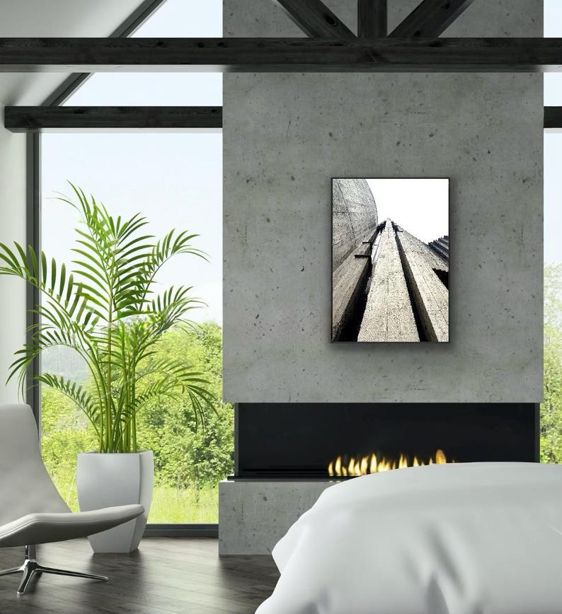 Op de afbeelding: Een rechthoekige wandkunstprint met een zwart-wit architectuurfoto van hoge, getextureerde betonstructuren die omhoog reiken naar een heldere lucht, weergegeven in een dunne zwarte lijst op een grijze betonnen openhaardmuur.