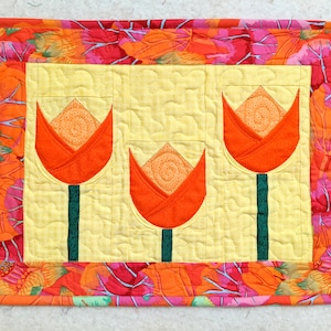 May include: A yellow quilted fabric wall hanging with a floral border and three orange fabric tulips.