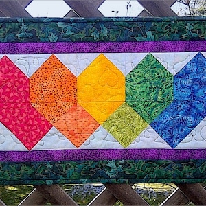 以下が含まれることがあります： 赤、オレンジ、黄色、緑、青のダイヤモンドの形をした虹色のパターンが施されたカラフルなキルトのテーブルランナー。ランナーは白い背景と紫と緑の縁取りがあります。