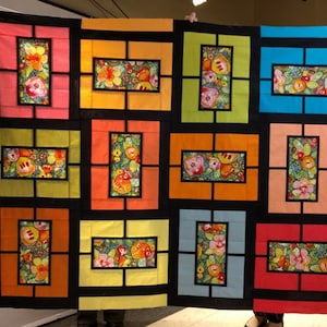 May include: A large, colourful quilt with a repeating pattern of rectangular blocks. Each block features a vibrant floral design within a contrasting frame. The quilt's background is black, and the blocks are in shades of pink, orange, yellow, green, and blue.