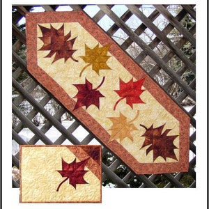 Pode incluir: Um corredor de mesa com fundo bege e borda marrom apresenta cinco folhas de bordo em tons de vermelho, laranja e amarelo. As folhas estão dispostas em diagonal. Também é mostrado um jogo americano correspondente com uma única folha de bordo vermelha.