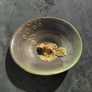 May include: A round ceramic bowl with a dark grey and green glaze. The interior features a raised, textured sculpture of a goldfish in the centre, surrounded by several small, circular embossed bubble patterns rising towards the rim.
